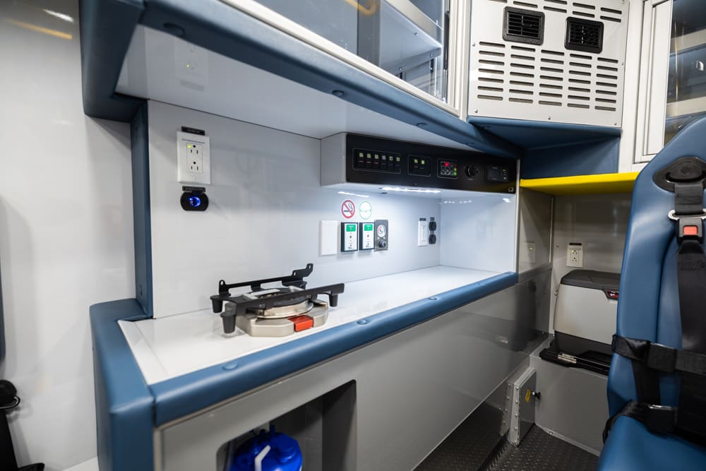 Demers ambulance patient compartment work counter with integrated equipment mount, electrical outlets and controls, under-cabinet LED task lighting, and overhead storage next to a forward-facing attendant seat
