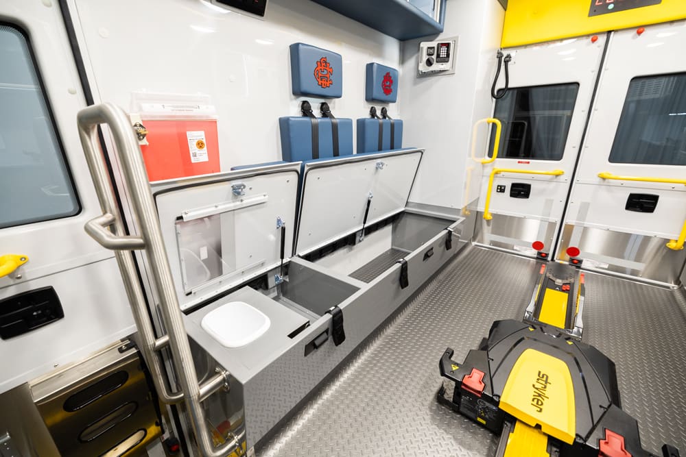Demers ambulance patient compartment bench with hinged under-seat storage compartments open, integrated waste container, rear attendant seating, and center-mounted stretcher loading system