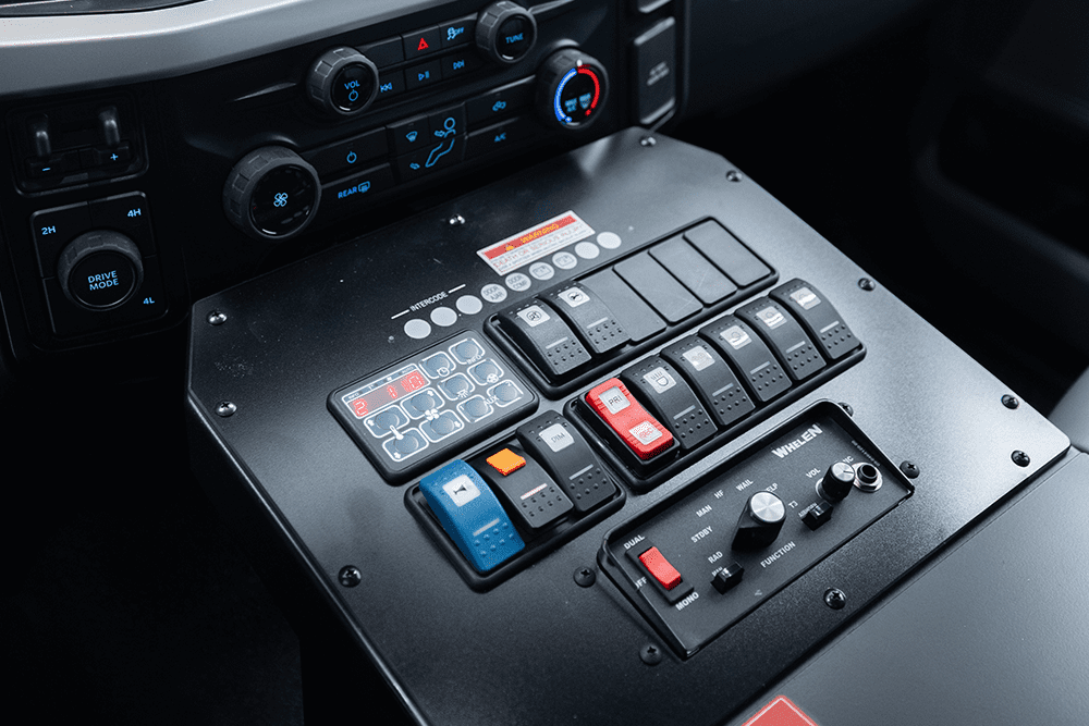 Close-up of a Demers ambulance center console switch panel, showing labeled rocker switches and control modules for emergency lighting, sirens, and vehicle systems positioned below the cab climate controls