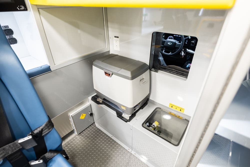 Demers ambulance interior featuring a securely mounted medical refrigerator beneath a work surface, with adjacent cab-to-module pass-through window and floor access panel for service and storage