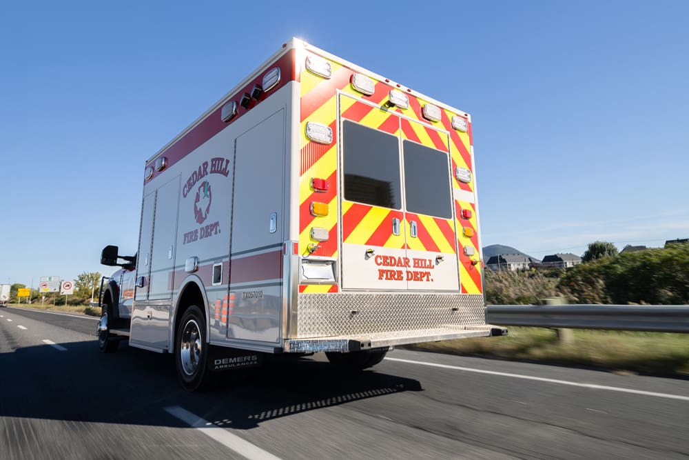 Rear three-quarter view of a Cedar Hill Fire Department Demers ambulance driving on a highway, featuring high-visibility red-and-yellow chevron striping and rear emergency lighting under clear blue skies
