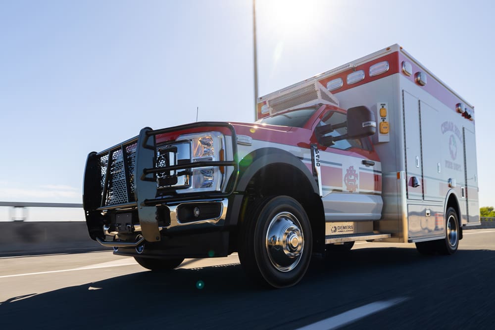 Cedar Hill Fire Department Demers ambulance driving on a highway in daylight, shown from a low front-side angle with emergency lighting and protective grille visible