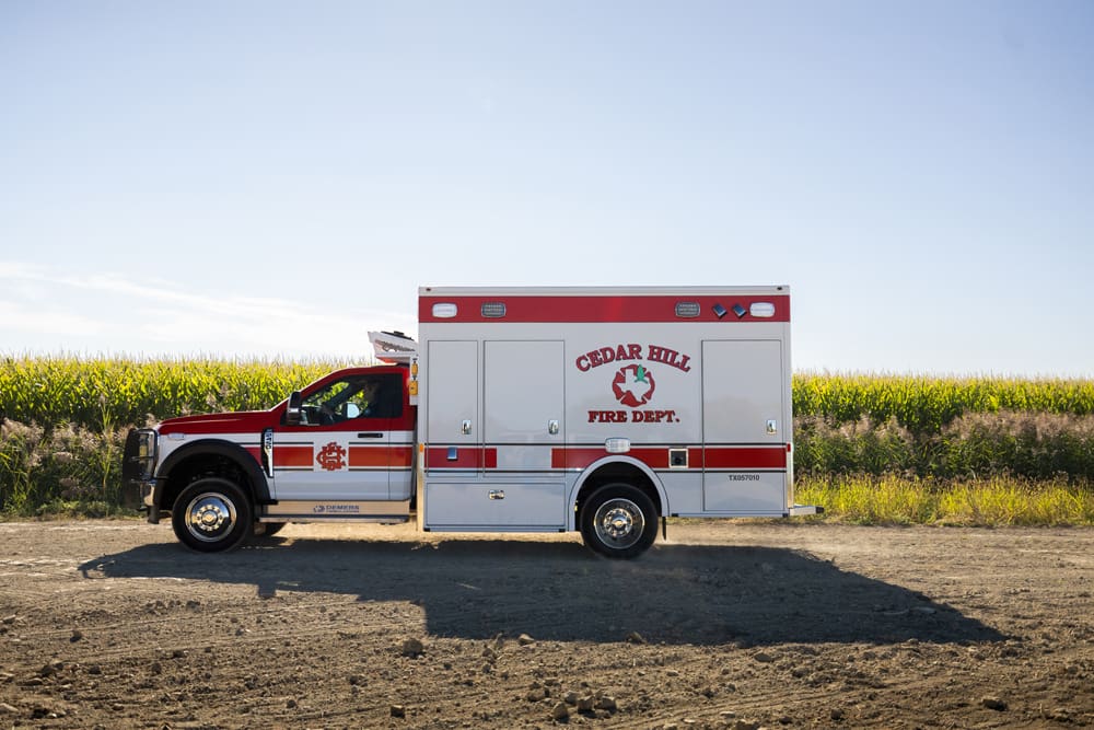 Cedar Hill Fire Department Demers ambulance shown in full side profile, parked on a rural dirt road beside a cornfield under clear daylight