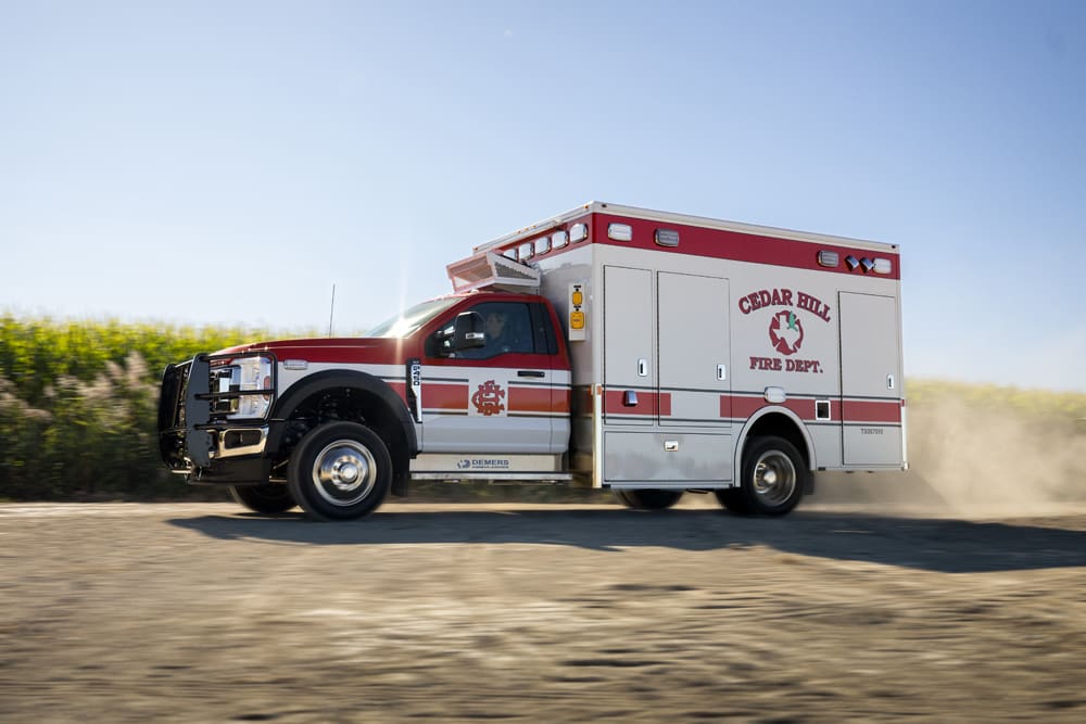 Side view of a Cedar Hill Fire Department Demers ambulance driving on a rural gravel road, kicking up dust with cornfields and a clear blue sky in the background