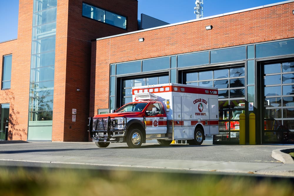 Cedar Hill Fire Department Demers ambulance parked outside a modern fire station with glass bay doors, shown in daylight from a wide exterior view