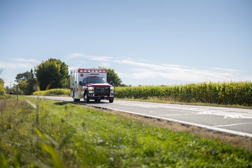 Front view of a Cedar Hill Fire Department Demers ambulance driving along a rural two-lane road, bordered by green fields and cornfields under a clear blue sky