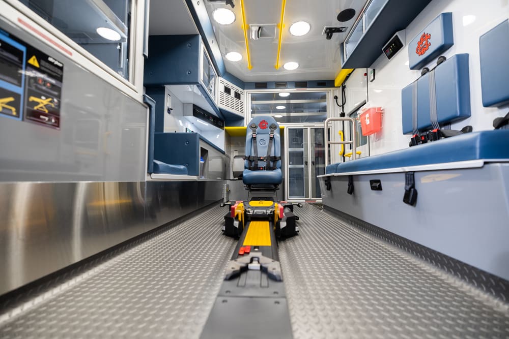 Centered view down the Demers ambulance patient compartment showing a powered stretcher loading track, forward-facing attendant seat, bench seating with secure restraints, glass-front storage cabinets, overhead grab rails, and bright LED lighting