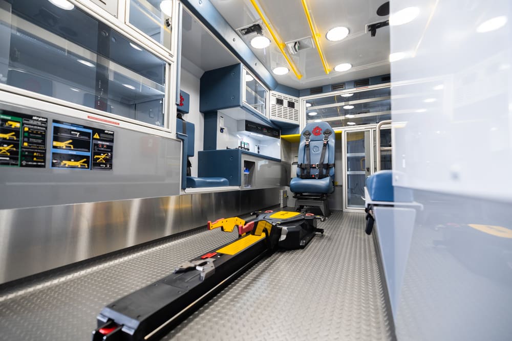 Interior view of a Demers ambulance patient compartment featuring a powered stretcher loading system on the floor, aluminum cabinetry with clear doors, integrated work surfaces, and a forward-facing attendant seat under bright LED ceiling lighting