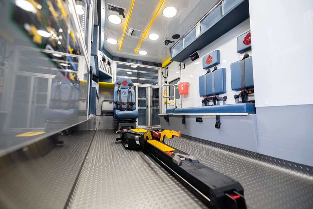 Interior view of a Demers ambulance patient compartment showing a powered stretcher loading system centered on the floor, forward-facing attendant seat, bench seating with restraint belts, aluminum cabinetry, and bright LED ceiling lighting