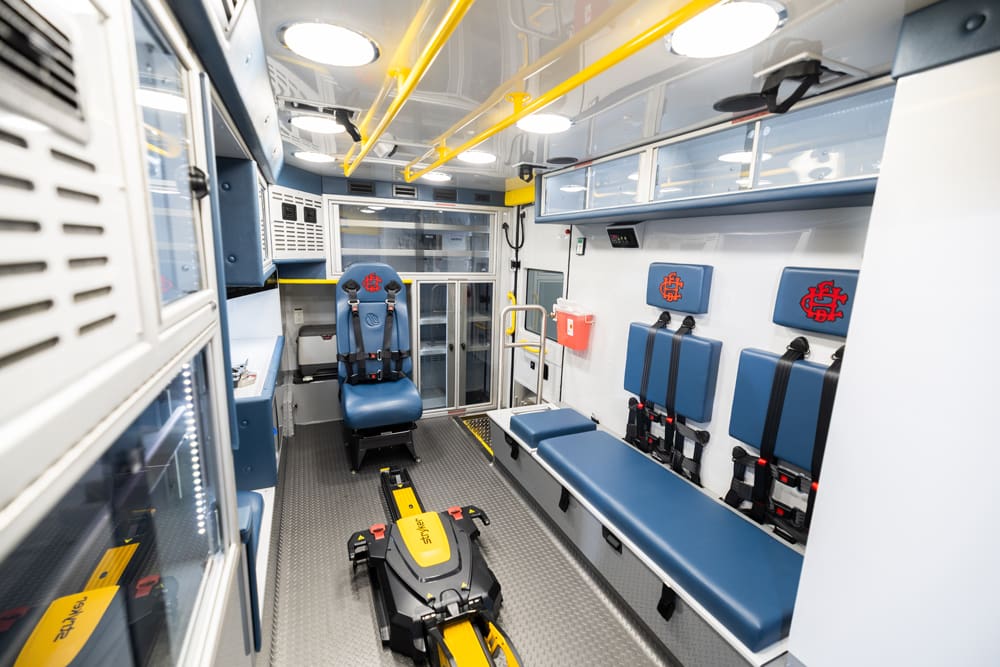 Wide interior view of a Demers ambulance patient compartment featuring a powered stretcher loading system, forward-facing attendant seat, bench seating with integrated restraints, overhead grab rails, glass-front cabinetry, and bright LED lighting