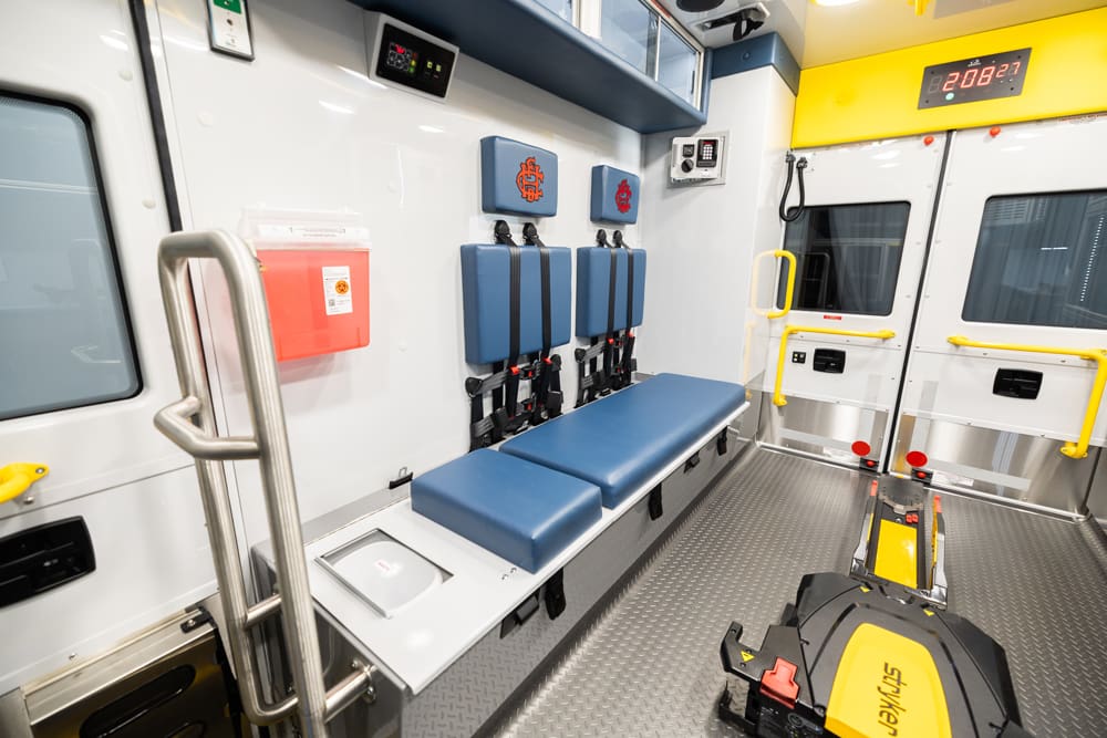 Demers ambulance patient compartment bench seating with blue cushions and integrated seatbelt restraints, wall-mounted sharps container, stainless grab rail, rear patient doors with yellow handles, and a powered stretcher loading system along the center aisle