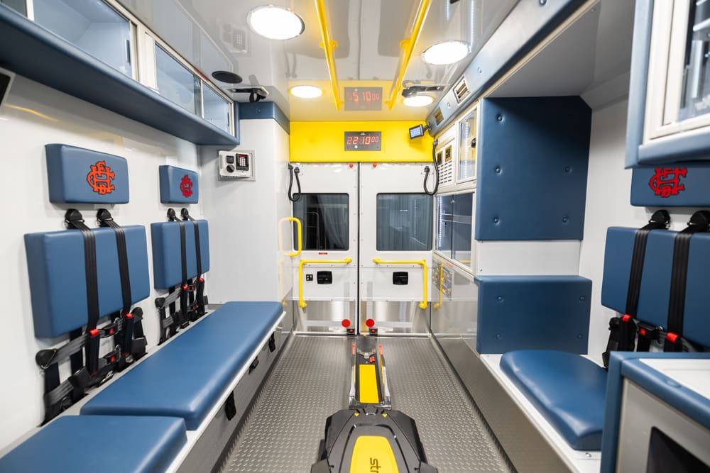 Wide interior view of the Demers ambulance patient compartment showing opposing bench seats with integrated restraints, a centered powered stretcher loading system, glass-front upper cabinets, bright LED ceiling lights, yellow grab rails, and rear patient doors