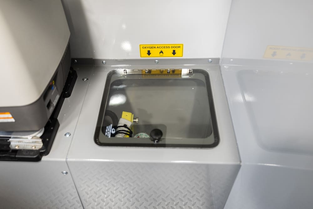 Close-up of a Demers ambulance oxygen access compartment with a clear viewing window, labeled access door, and visible oxygen fittings for quick inspection and service access