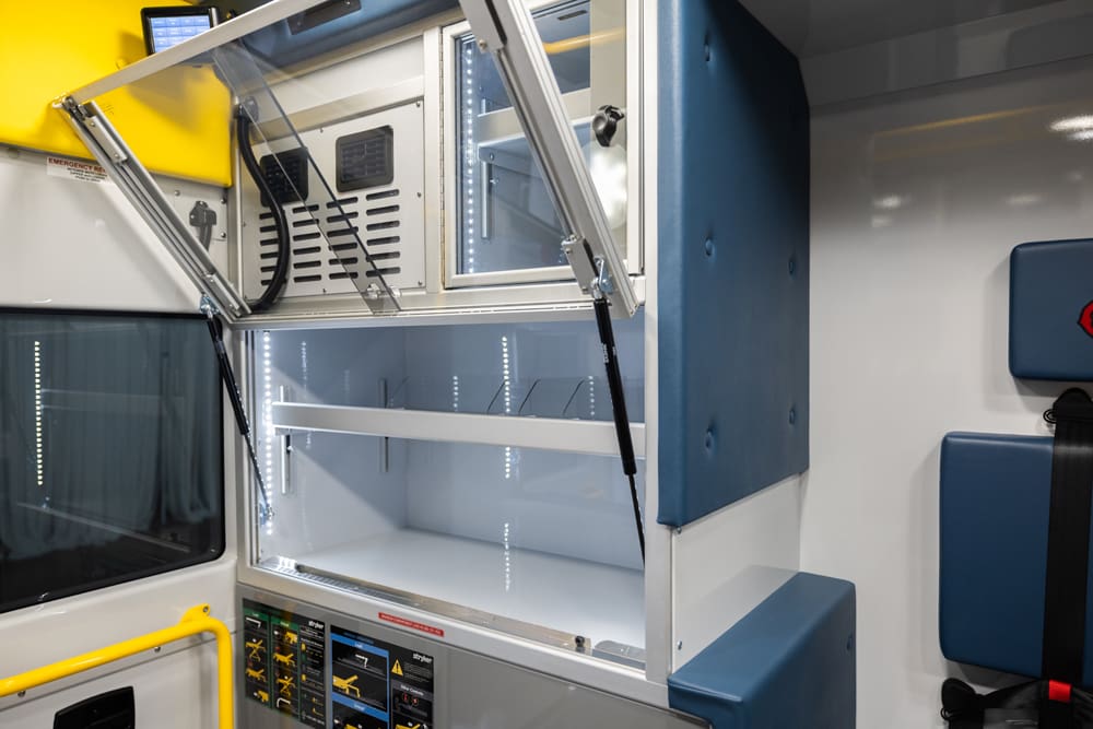 Interior Demers ambulance cabinetry with an open, upward-hinged clear cabinet door, showing illuminated storage shelves and integrated ventilation above the work surface in the patient compartment
