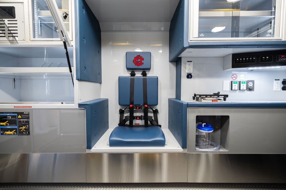 Forward-facing EMS attendant seat with integrated restraints, positioned between illuminated clear cabinetry and a patient-care work surface inside the Demers ambulance module