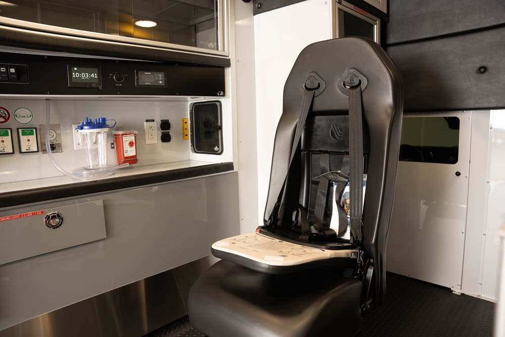 Interior view of a Demers ambulance patient compartment showing a rear-facing attendant seat with integrated restraints positioned beside the primary medical control panel, suction canister, and power outlets for in-transport care