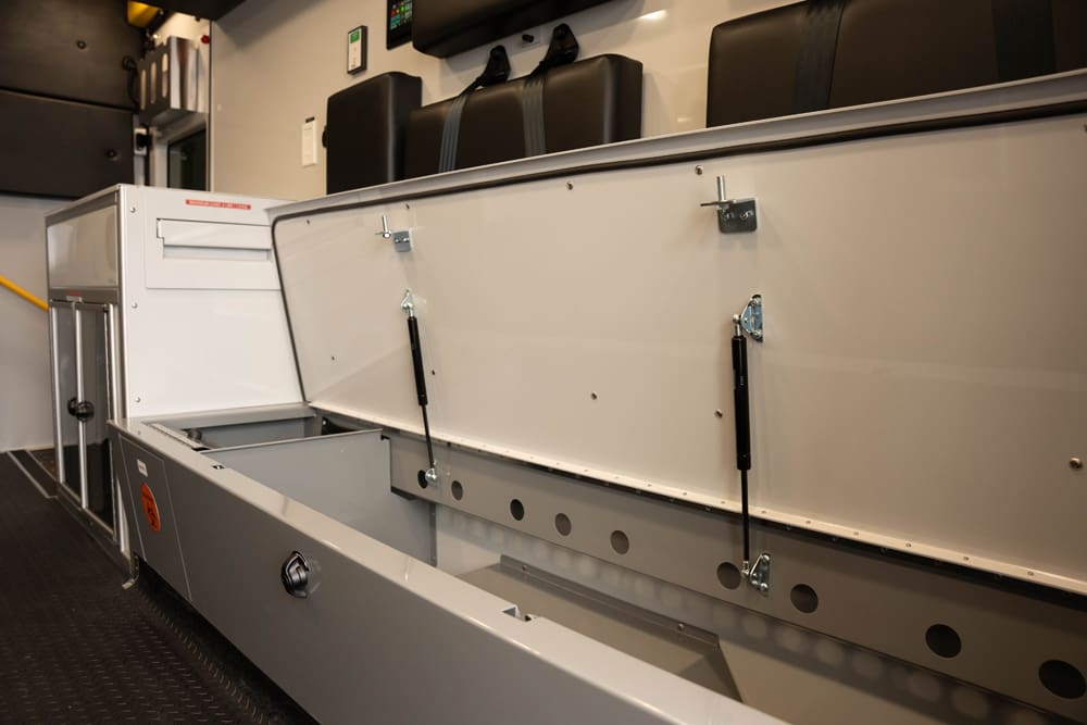 Open bench seat storage inside a Demers ambulance patient compartment, showing a lift-up lid with gas struts and a deep, integrated storage cavity designed for secure, easy-access equipment storage during transport