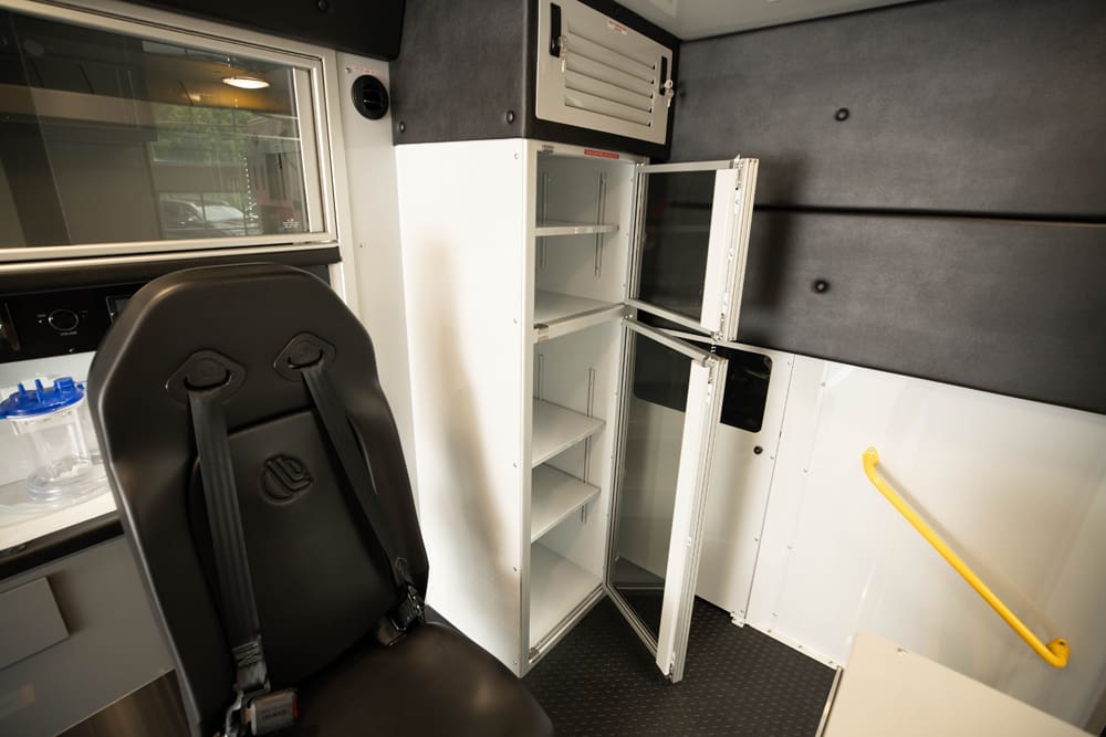 Interior view of a Demers ambulance patient compartment showing a secured attendant seat next to a tall, glass-front storage cabinet with open doors and adjustable shelving for organized medical supplies
