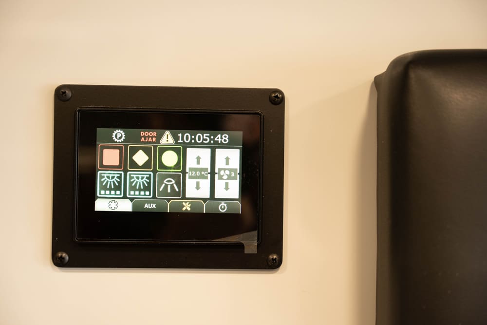 Touchscreen multiplex control panel inside a Demers ambulance, displaying system status icons, lighting controls, HVAC settings, and door alerts for centralized vehicle management