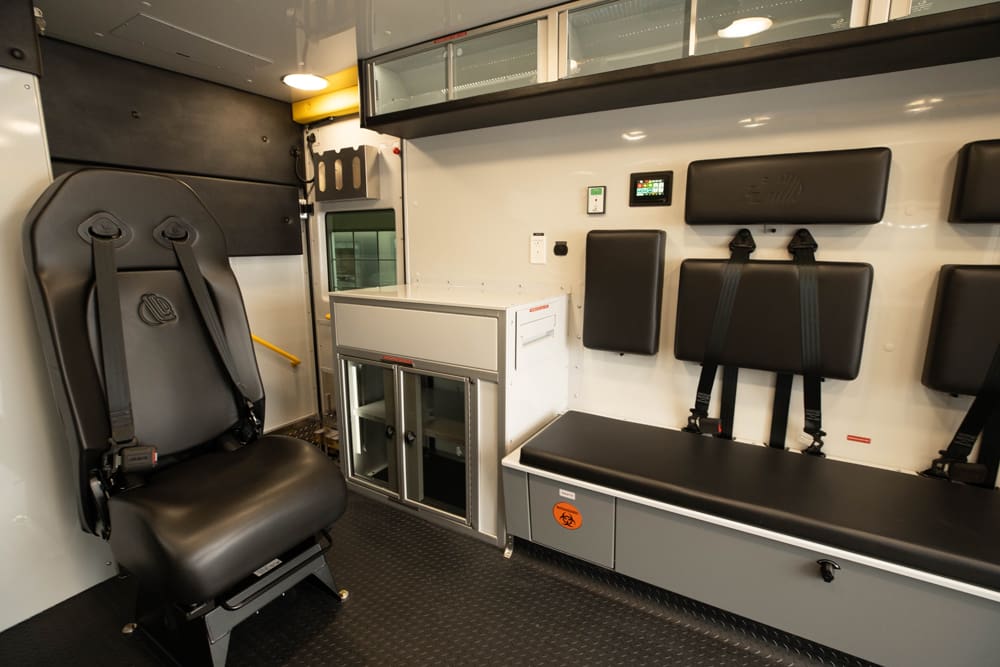 Interior view of a Demers ambulance patient compartment featuring a forward-facing attendant seat, bench seating with integrated restraints, glass-front cabinetry, and a compact work surface designed for organized, in-motion patient care