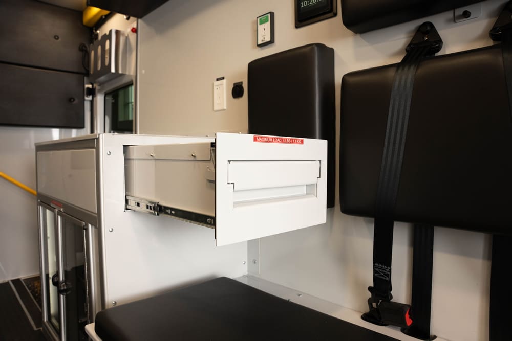 Close-up of a Demers ambulance patient compartment showing a slide-out storage drawer integrated beneath bench seating, with secure restraints and adjacent work surfaces designed for organized, in-motion access to supplies