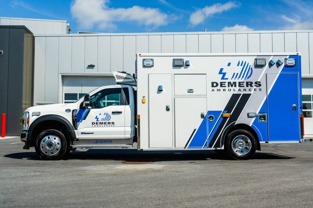 Side profile of a Demers ambulance with blue-and-white graphics parked outside a modern service facility, highlighting the full-length patient module, exterior compartments, and Ford chassis
