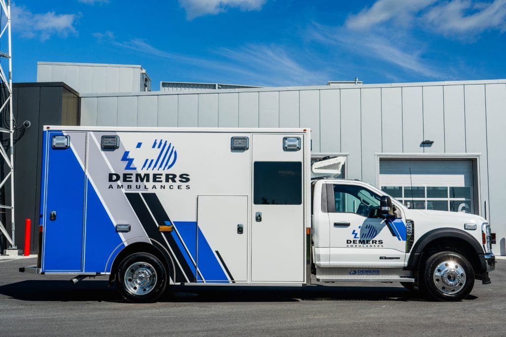 Side profile of a Demers ambulance parked outside a modern service facility, featuring blue-and-white branding, exterior compartment doors, emergency lighting, and a Ford chassis under a clear blue sky