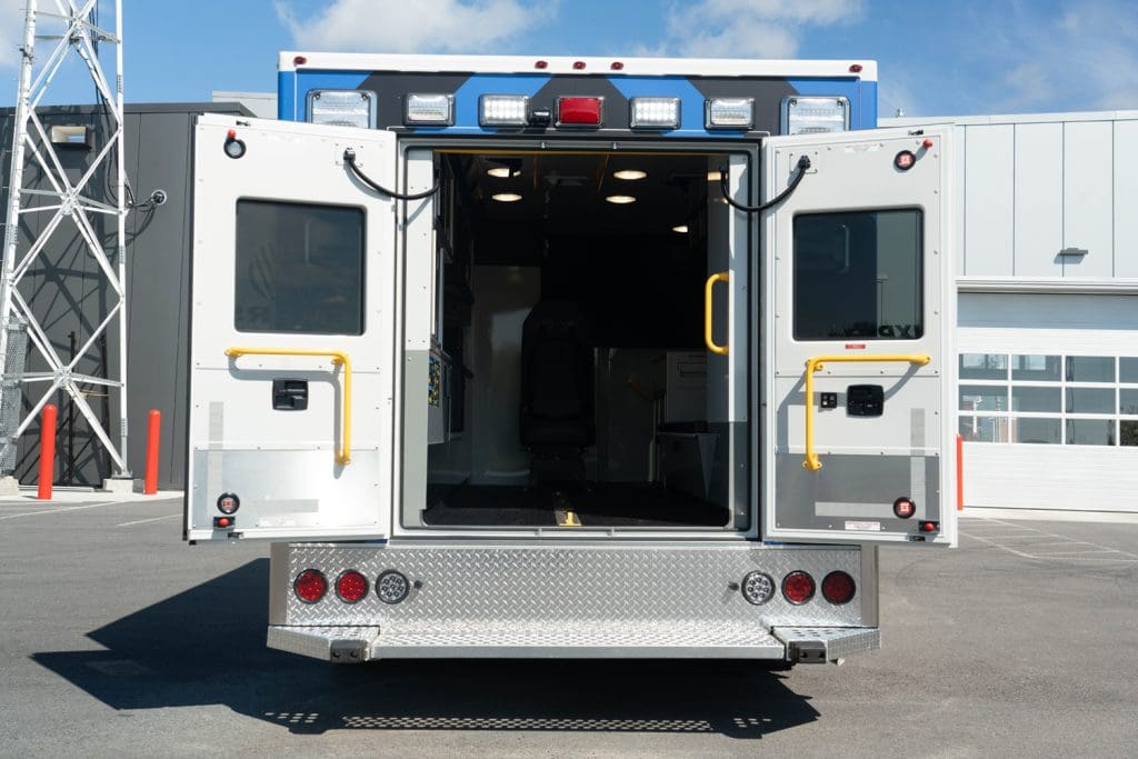 Rear view of a Demers ambulance with both rear patient compartment doors open, showing the illuminated interior entry, high-visibility grab handles, exterior lighting, and rear step at a service facility