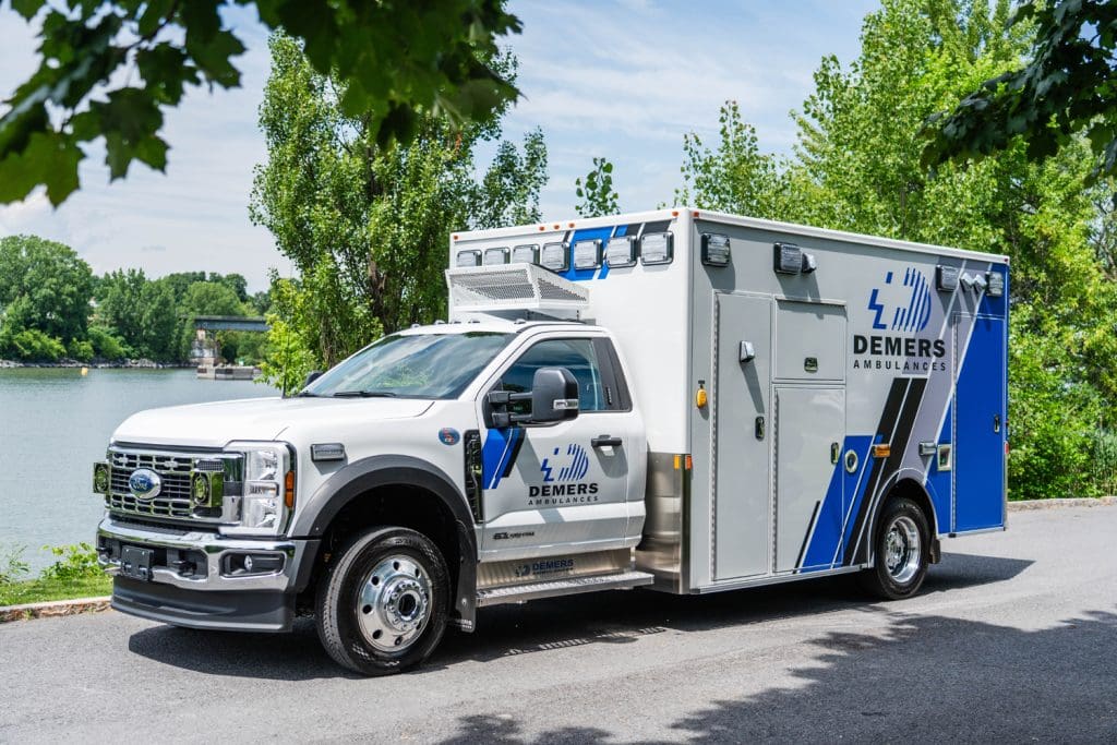 White Demers ambulance with blue branding parked along a riverside road, showing the exterior patient module, side compartments, and Ford chassis in a natural, tree-lined setting