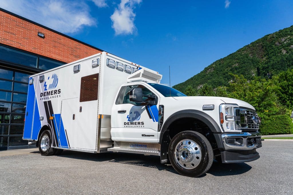 Demers ambulance with blue-and-white graphics parked outside a modern facility, showing the front three-quarter view, Ford chassis, exterior compartments, and mountain landscape in the background