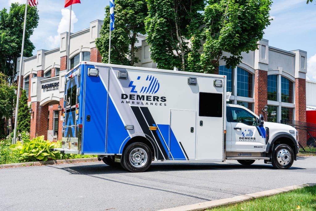 Demers ambulance with blue-and-white branding parked outside the Demers Ambulances facility, showing the side profile, exterior compartments, and Ford chassis against a modern brick building backdrop