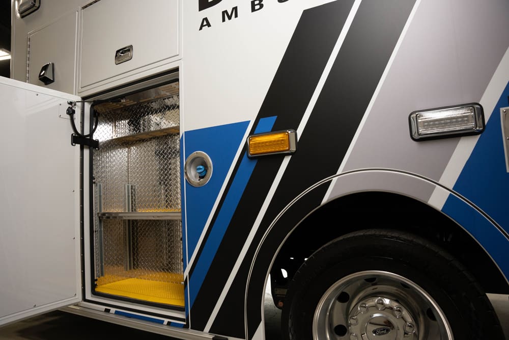 Close-up of a Demers ambulance exterior showing an open side storage compartment with diamond-plate interior shelving, adjacent wheel well, and blue-and-black graphic striping on the patient module