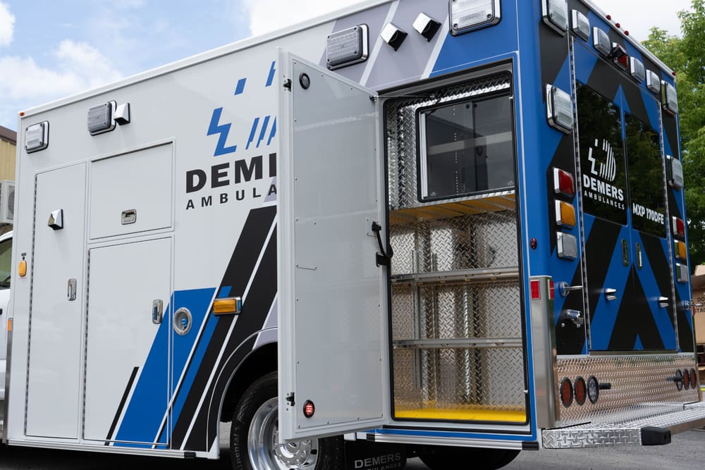 Rear-side view of a Demers ambulance with an open exterior storage compartment, showing diamond-plate shelving and interior lighting, with Demers branding and blue-and-black graphics on the patient module
