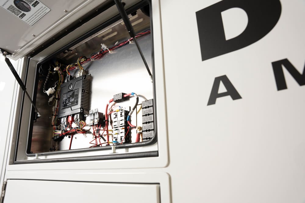 Open exterior electrical compartment on a Demers ambulance, showing organized wiring, control modules, and fuse blocks mounted inside the service-access panel