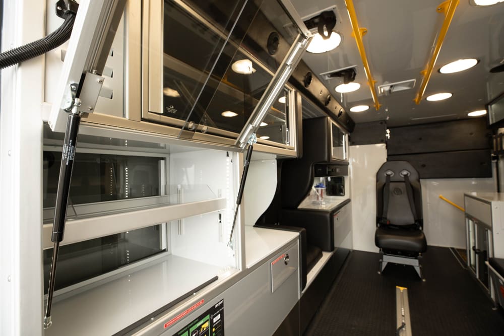 Interior view of a Demers ambulance showing open, glass-front overhead cabinets with gas-assist supports, illuminated patient compartment lighting, and a forward-facing attendant seat designed for organized, in-reach equipment access