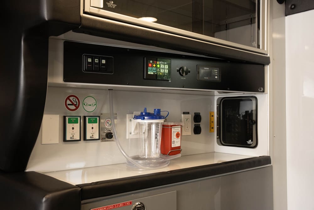 Close-up of a Demers ambulance patient care control panel with integrated switches, digital displays, suction canister, sharps container, and power outlets mounted above a clean work surface