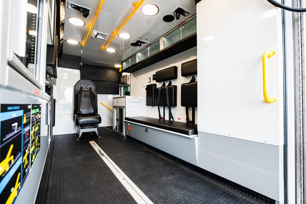 Interior view of a Demers ambulance patient module showing a forward-facing attendant seat, side bench with integrated restraints, overhead grab rails, glass-front cabinets, and open center aisle