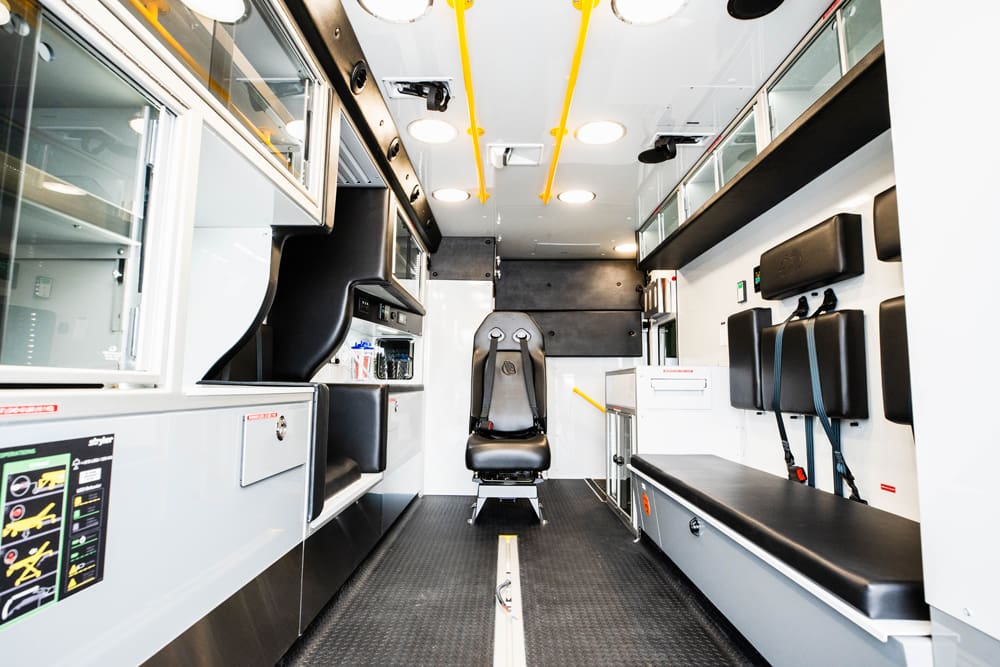Interior view of a Demers ambulance patient module featuring a centrally mounted attendant seat, rear-facing bench with integrated restraints, overhead grab rails, glass-front cabinetry, and streamlined storage