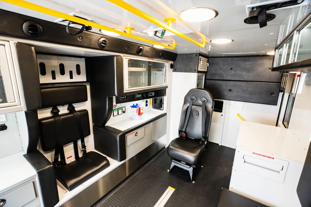 Interior view of a Demers ambulance patient module showing a forward-facing attendant seat, rear-facing bench seat with restraints, overhead grab rails, integrated cabinetry, and organized medical work surfaces