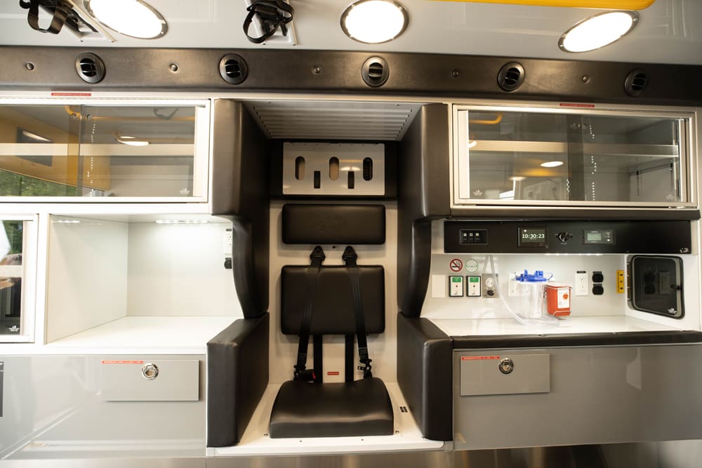 Interior view of a Demers ambulance patient module showing a centered, forward-facing attendant seat with integrated restraints, flanked by glass-front overhead cabinets, a work surface with medical controls, and overhead lighting and ventilation
