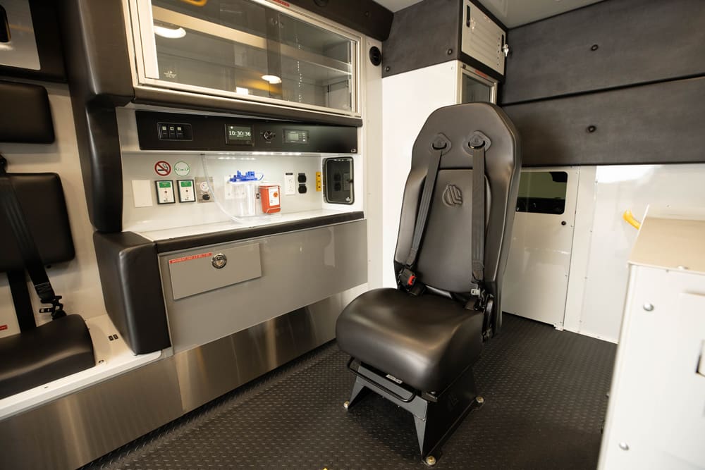 Interior view of a Demers ambulance patient module featuring a forward-facing attendant seat positioned beside a side work counter with integrated medical controls, suction canister, power outlets, and glass-front overhead storage