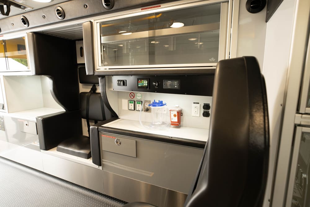 Interior view of a Demers ambulance patient module showing a side work counter with integrated medical controls, suction canister, power outlets, and storage drawer beneath glass-front overhead cabinetry, with an attendant seat positioned for in-motion access