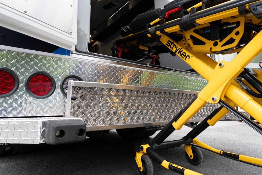 Rear loading area of a Demers ambulance with a yellow Stryker stretcher positioned at the open rear doors, showing the loading deck, step, and rear lighting details