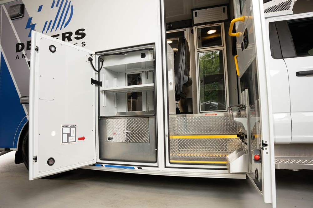 Exterior side access view of a Demers ambulance with the curbside compartment door open, showing organized interior storage shelves, battery layout labeling, and diamond-plate step entry into the patient module