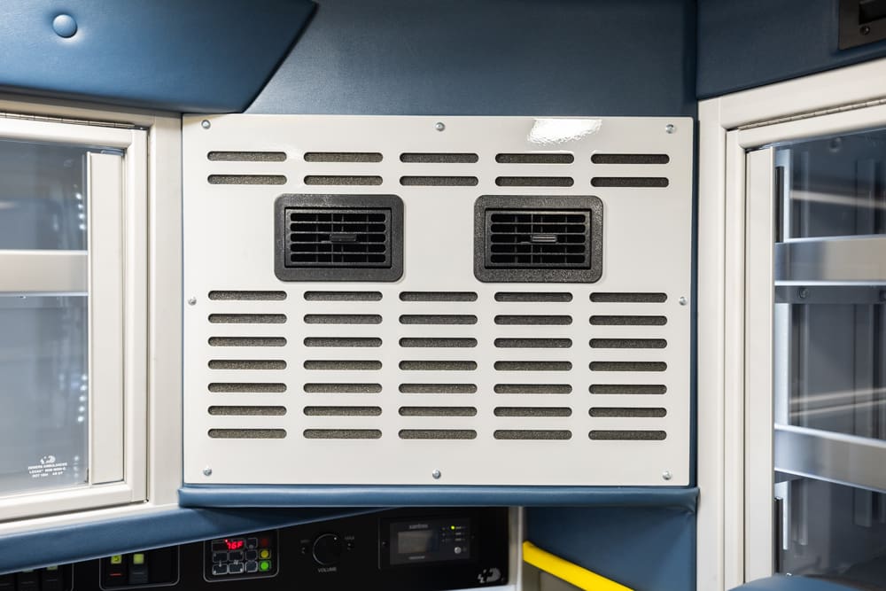 Close-up of the Demers ambulance patient compartment ventilation panel with dual air vents and perforated grille, positioned between glass-front storage cabinets for controlled airflow