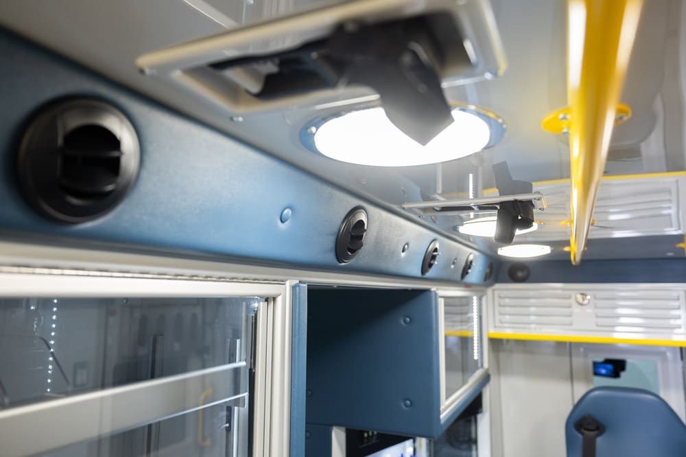 Close-up of the Demers ambulance patient compartment ceiling showing integrated LED dome lights, adjustable air vents, overhead cabinetry, and yellow grab rails for safety and accessibility