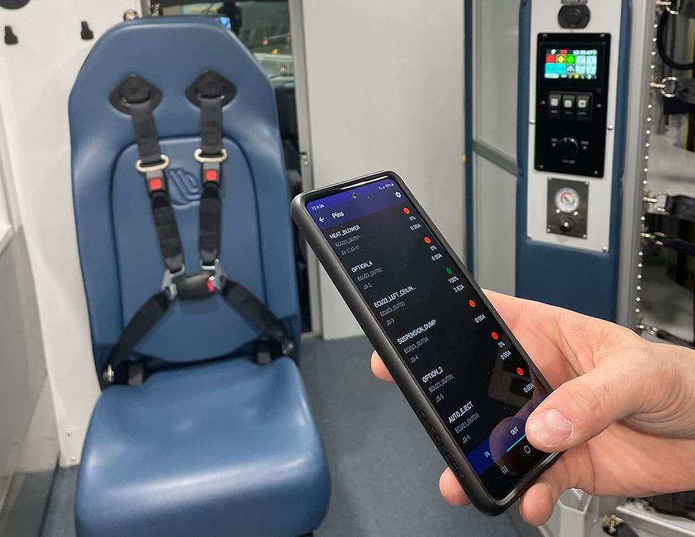 Hand holding a smartphone displaying Demers ambulance electrical system controls, with a blue attendant seat and interior control panel visible in the background
