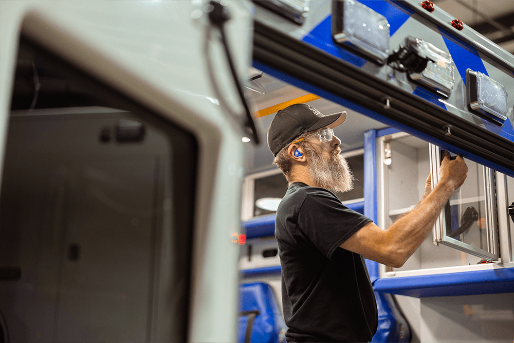 Demers ambulance manufacturing technician wearing safety glasses and hearing protection installs an exterior compartment door on a vehicle in the production facility