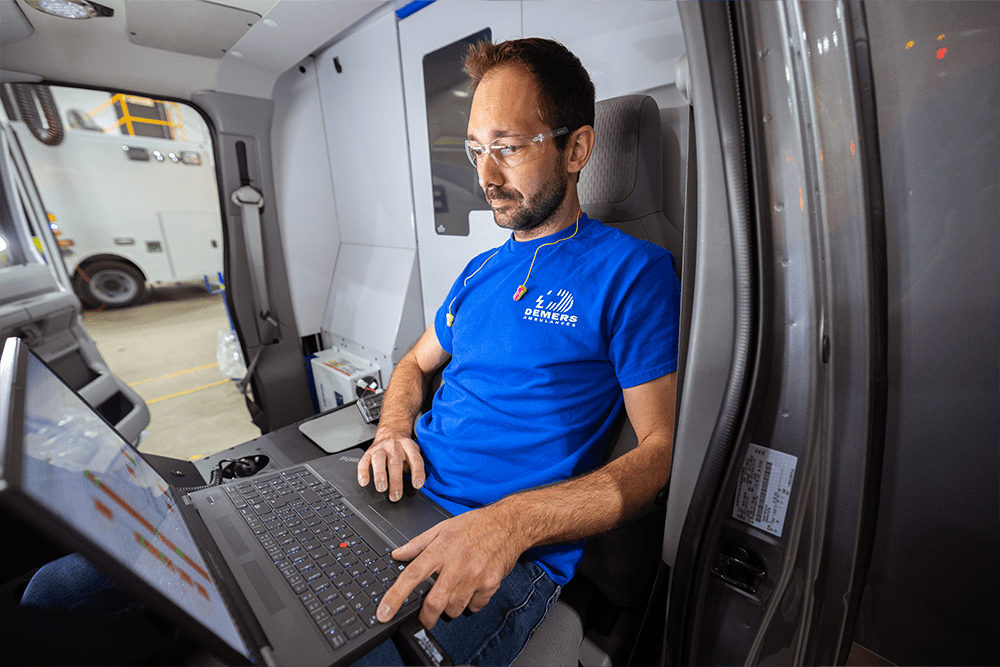 Demers Ambulances technician seated inside an ambulance cab, wearing safety glasses and a blue company shirt, working on a laptop during vehicle testing or configuration inside the manufacturing facility