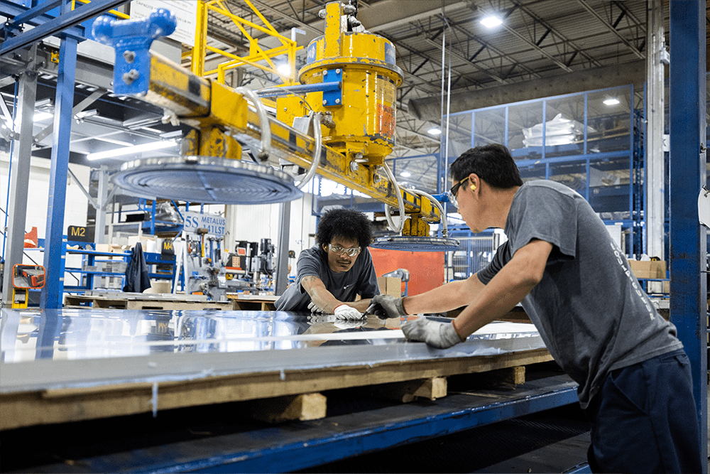 Two Demers manufacturing technicians wearing gloves and safety glasses work together shaping a large aluminum panel using industrial equipment inside an ambulance production facility